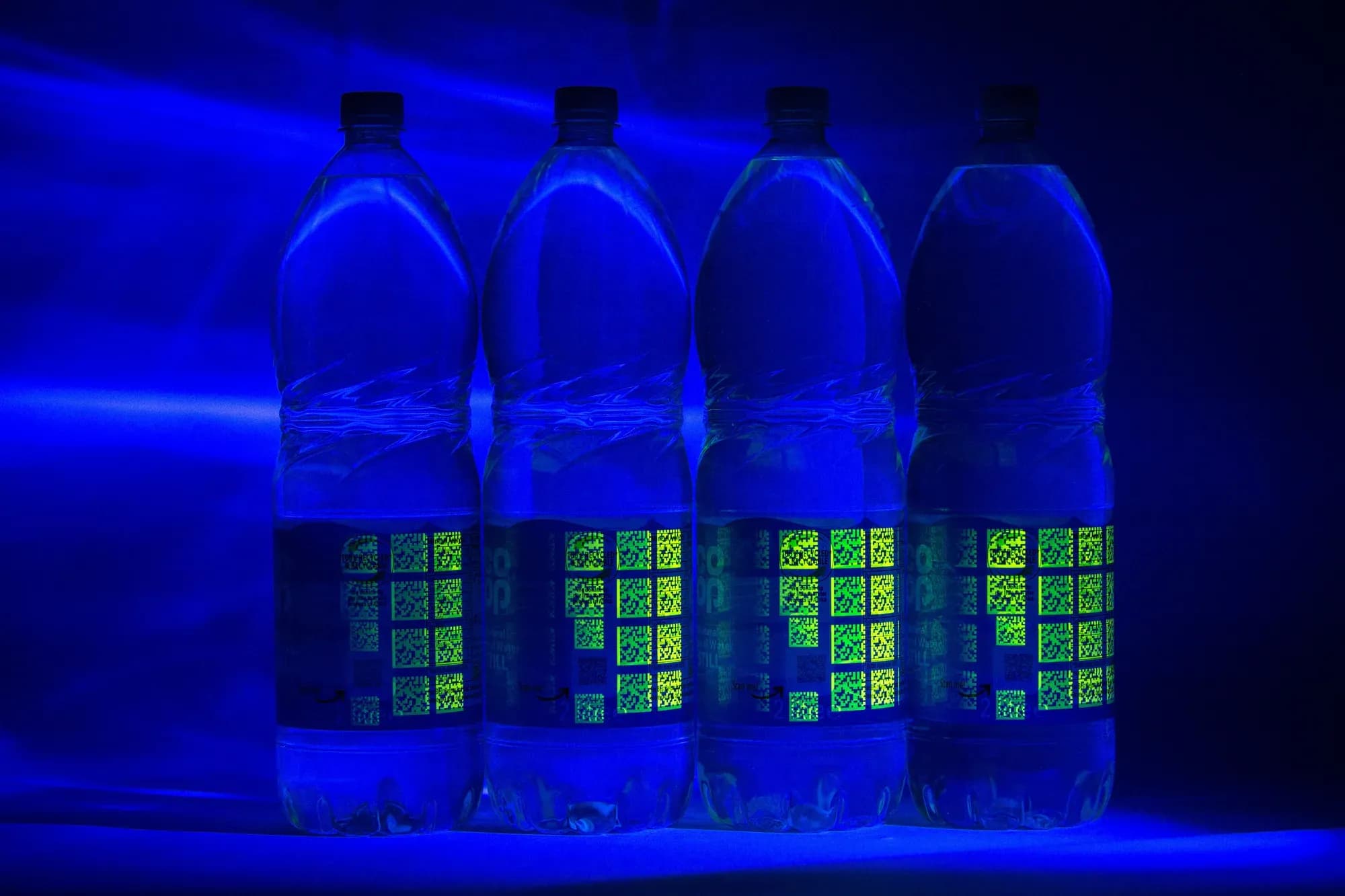 Four Co-op water bottles under UV light, the Invisible UV Tags glowing across their labels