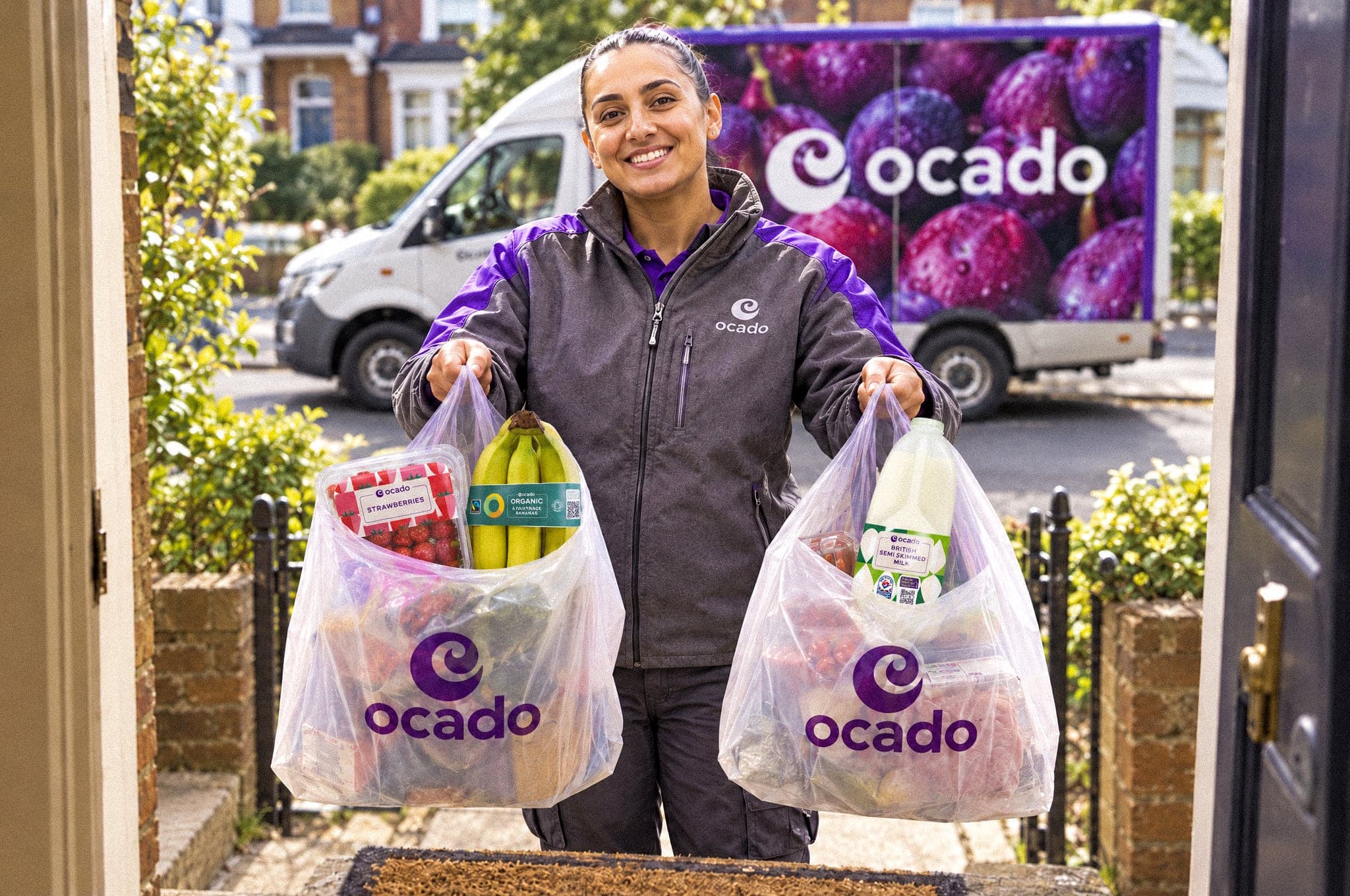 An Ocado delivery driver at a front door holding up two bags of Ocado shopping, including a bottle of Ocado milk, with an Ocado van parked behind